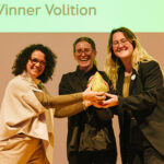 Magdalena Jo Umkehrer and Jana Rowenski standing on stage with jurymember Raquel Noronha receiving the iphiGenia trophy