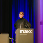 Luisa Nickolaus standing on stage behind a speakers desk in the MAKK.