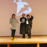 Petra Eisele and Isabel Naegele on stage with Claudia Herling while receiving the iphiGenia trophy