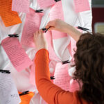 A woman hanging up pink an orrange notes onto a blackboard with other notes of the same kind.