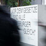 Text wall saying: All citizens are equal before the law. Before an behind the wall there are shadows of moving people.