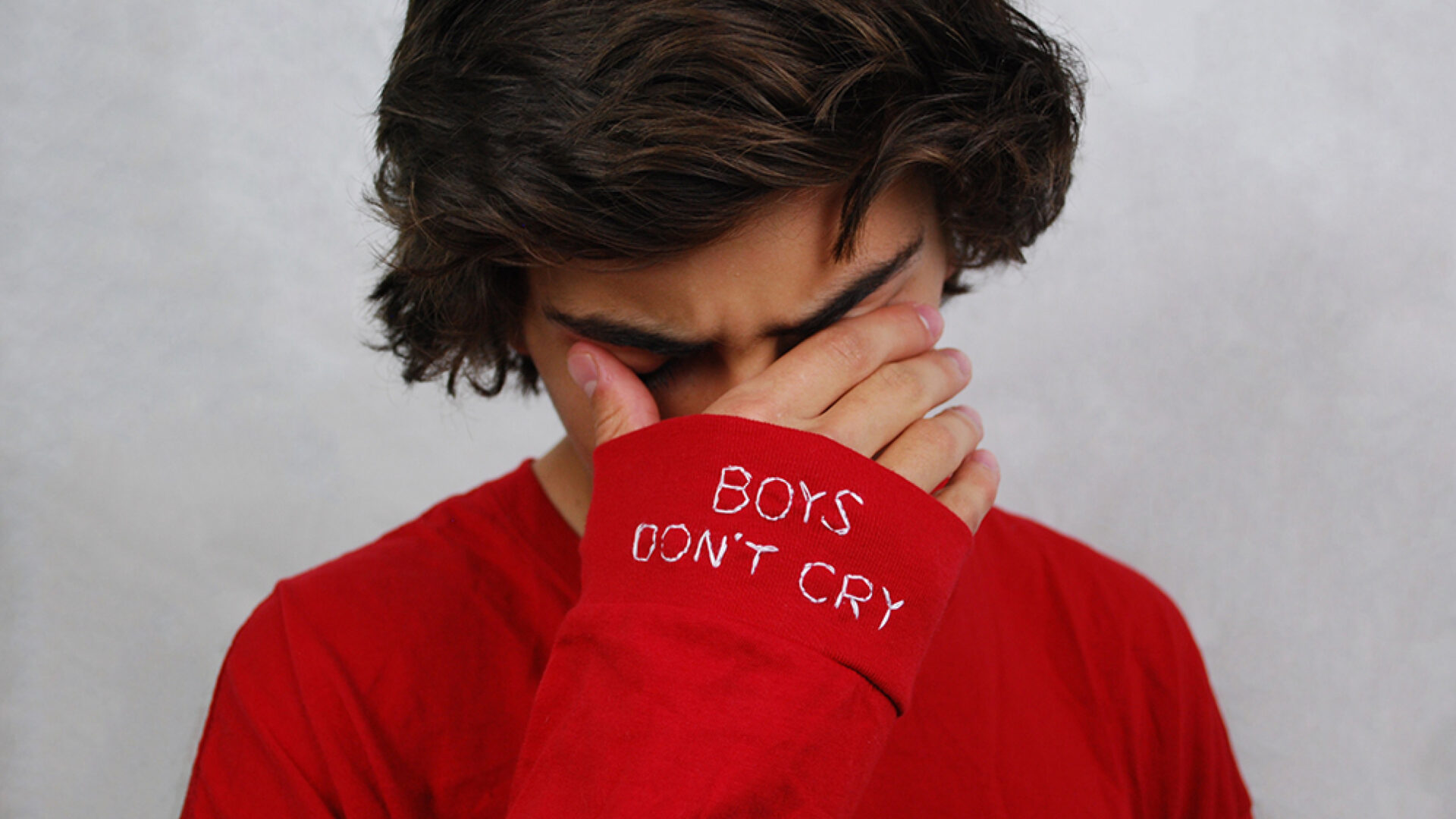 young man, covering his eyes with his hands. on the sleave of his red shirt is written 